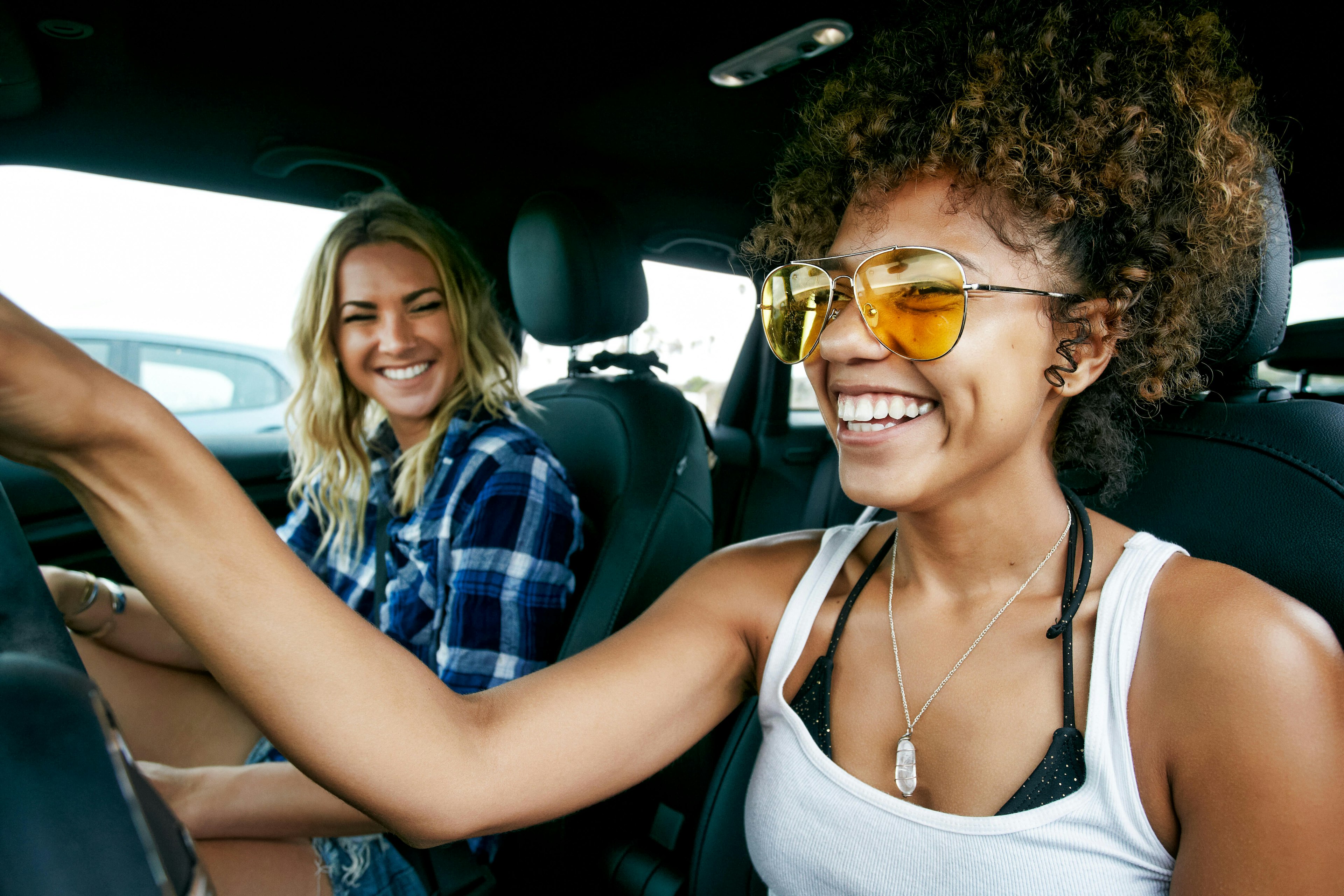 Portrait of two women with long blond and brown curly hair sitting in car, wearing sunglasses, smiling. - stock photo