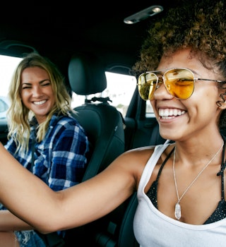 Portrait of two women with long blond and brown curly hair sitting in car, wearing sunglasses, smiling. - stock photo