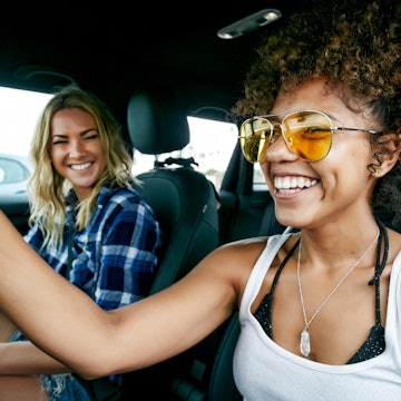 Portrait of two women with long blond and brown curly hair sitting in car, wearing sunglasses, smiling. - stock photo