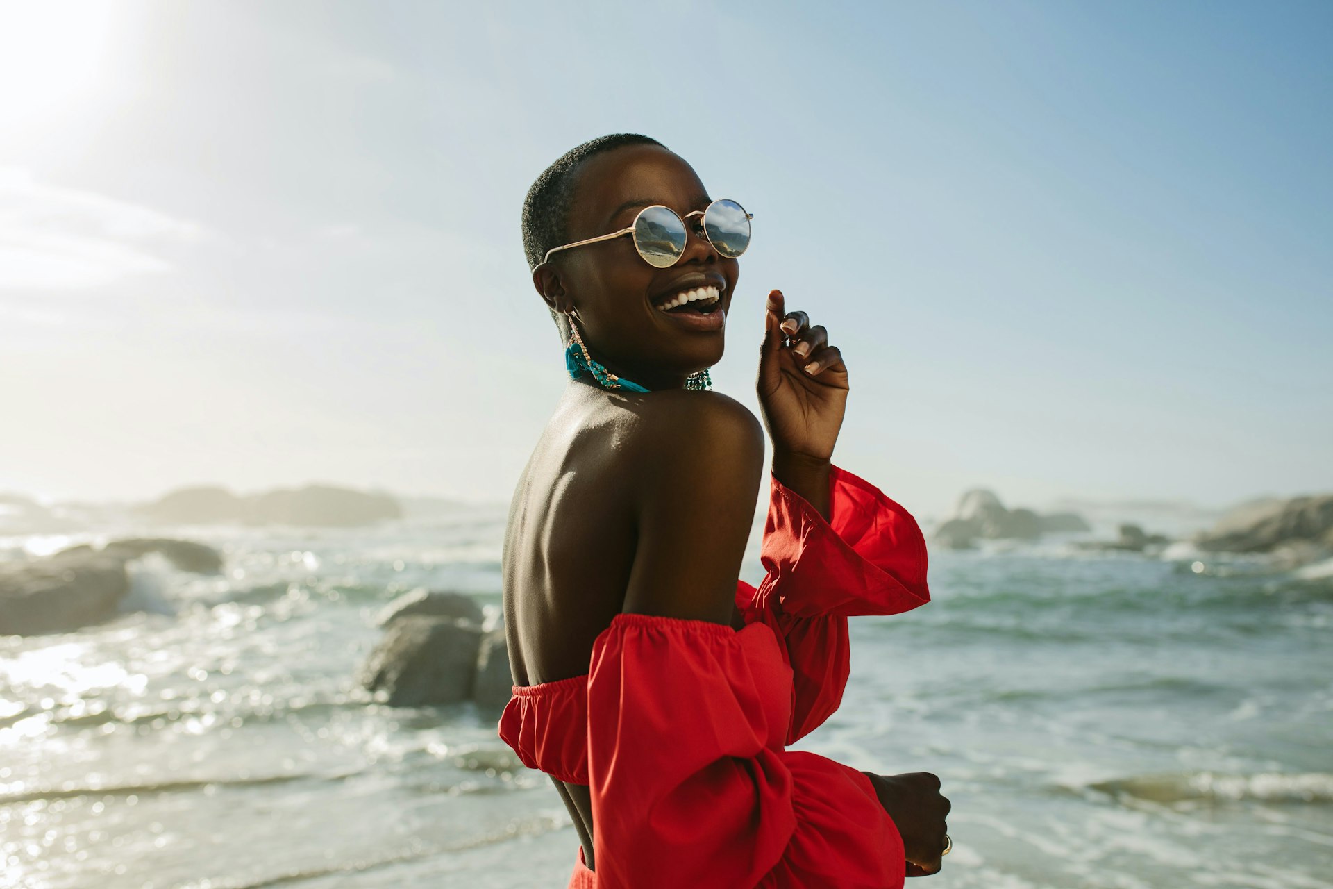 A black woman wearing a red dress and sunglasses looks over her shoulder and laughs while on the beach