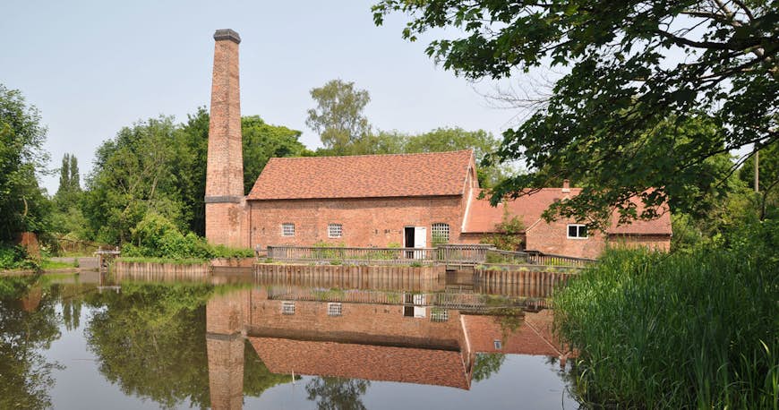A large brick mill building with a tall chimney on the edge of a pond