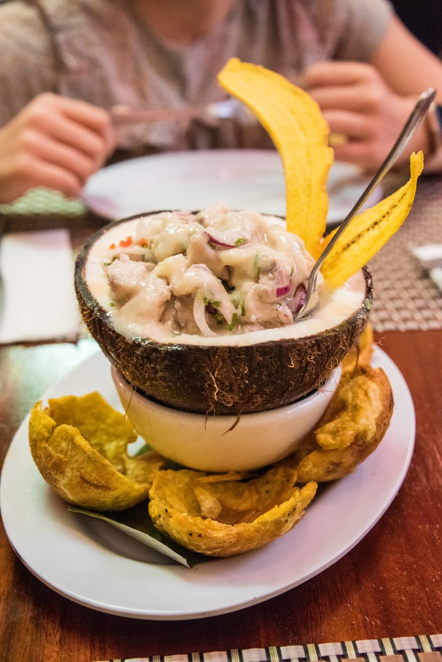 Poisson blanc tropical mariné dans du jus de citron et des oignons hachés, servi dans une noix de coco, des paniers de plantain vert entourant l'assiette.