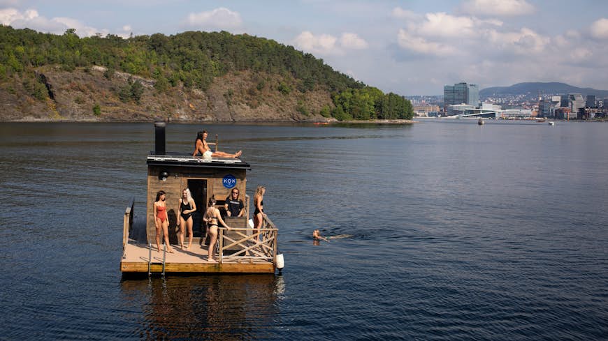 People use the KOK floating sauna in the fjord next to Oslo.  One was swimming, another of him was sunbathing on the roof, the other was standing on the deck. The driver is wearing a black T-shirt.  