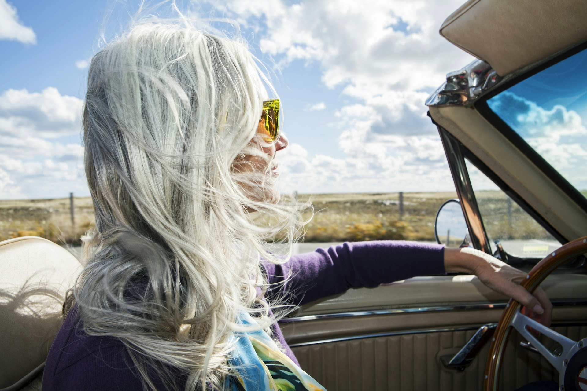 Woman from the baby boomer generation driving in an antique convertible car on a sunny fall day with her long, silver gray hair blowing in the wind