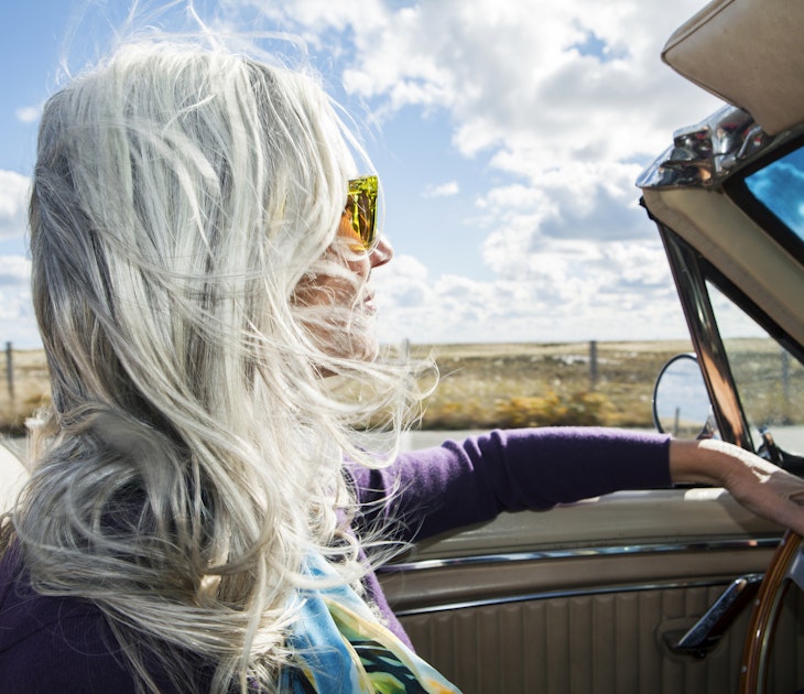 Woman from the baby boomer generation driving in an antique convertible car on a sunny fall day with her long, silver gray hair blowing in the wind.