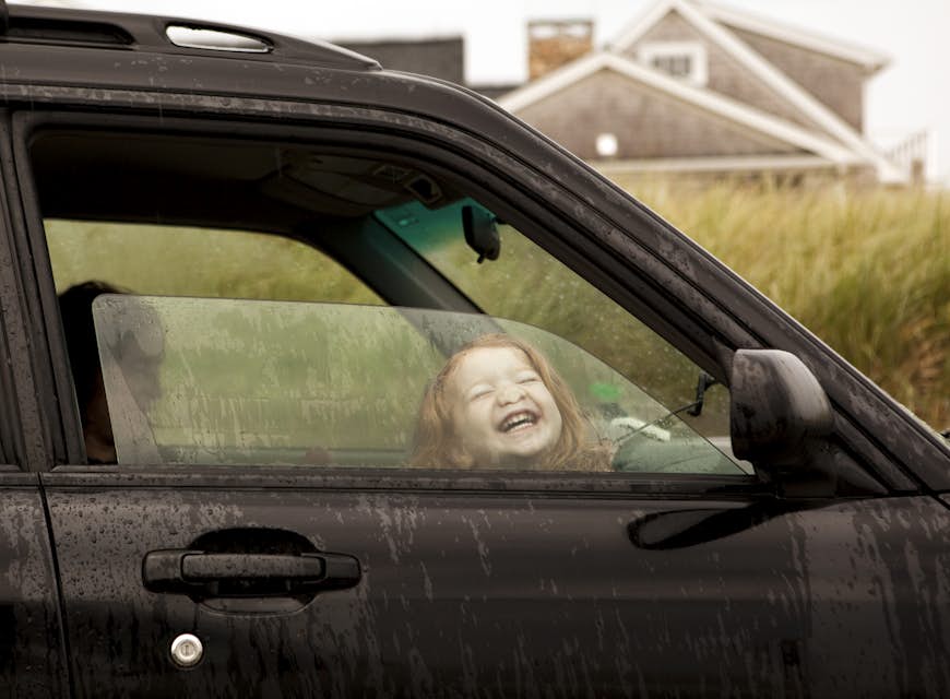 ingefärshårig vit flicka, två eller tre år gammal, trycker sitt ansikte mot ett halvöppet bilfönster i en blöt Cape Cod