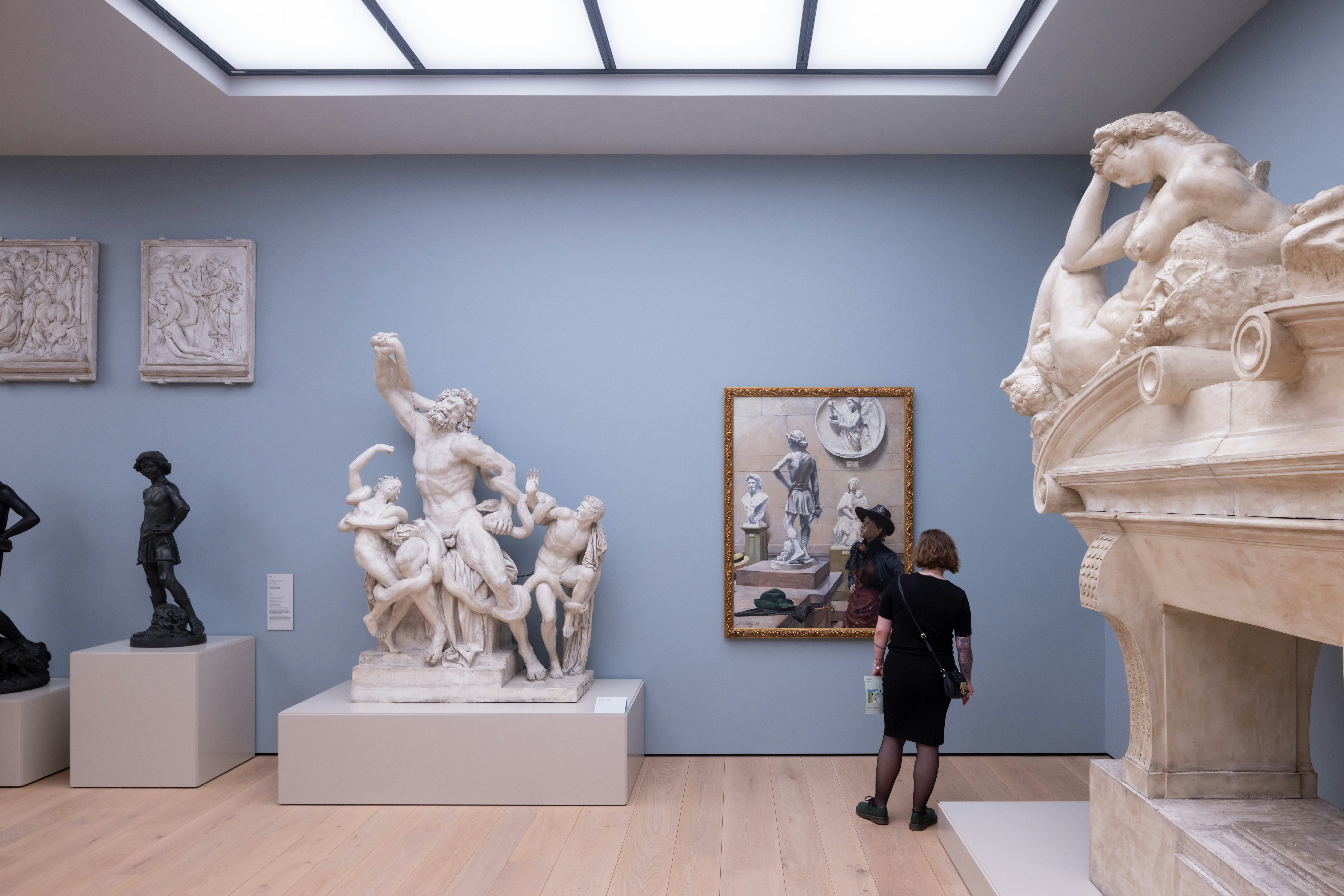 A white woman looks at an artwork at the National Museum of Norway in Oslo. We see her from behind. She is looking at a painting. There are also statues and pale blue walls.