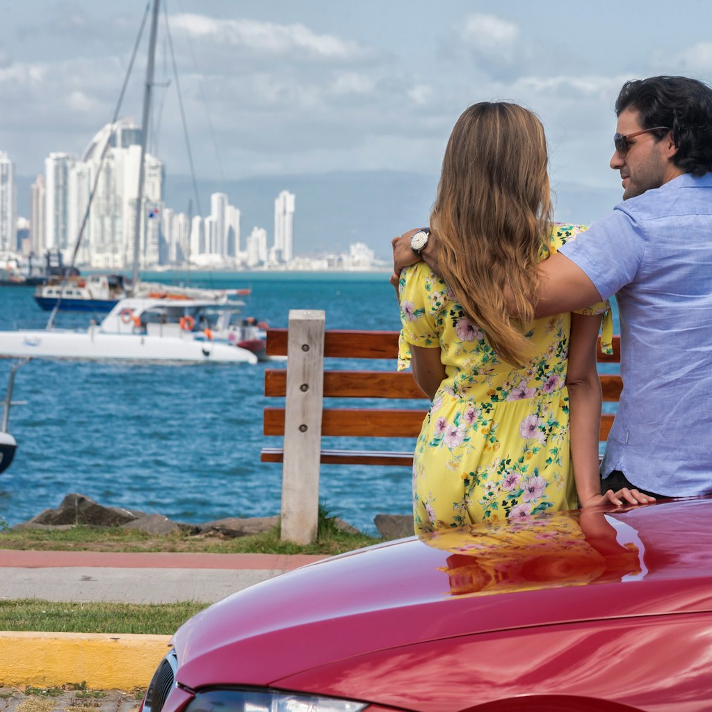 Latin Couple leaning on the side of a red car watching Panama city