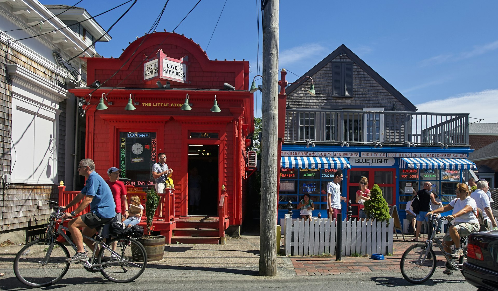A couple rides their bikes past two busy stores on a sunny day in Provincetown, Mass.