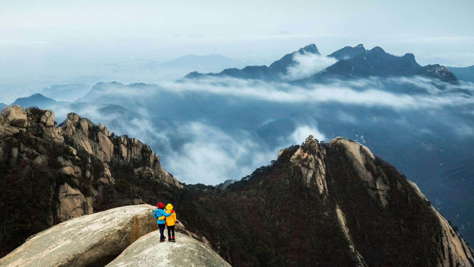 An aerial view of two hikers on a rocky outcrop overlooking cloudy mountain peaks at Bukhansan National Park, Seoul, South Korea