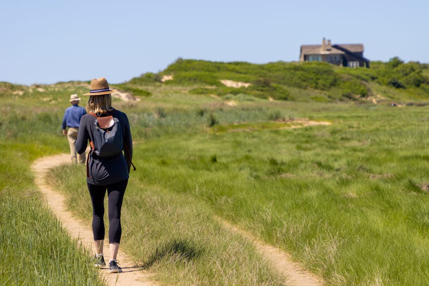 A woman hikes through a coastal area surrounded by grassy sand dunes