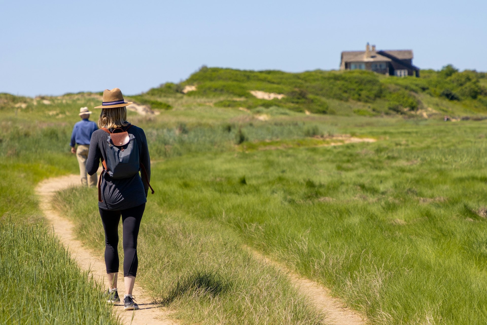 A woman hikes through a coastal area surrounded by grass-topped sand dunes