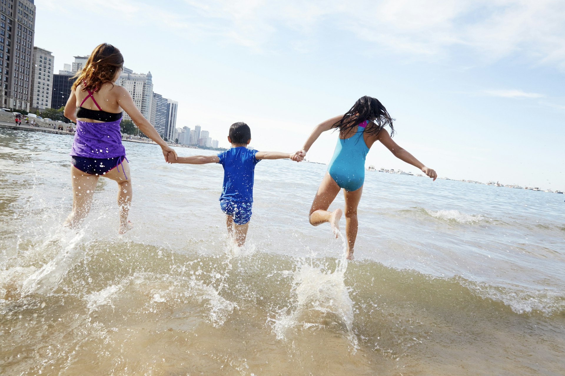 Three children hold hands as they run into the water of Lake Michigan with towers in the distance at a city beach in Chicago, Illinois, Midwest, USA