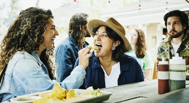 A laughing woman feeds her partner a tortilla chip heaped with guacamole at a food truck