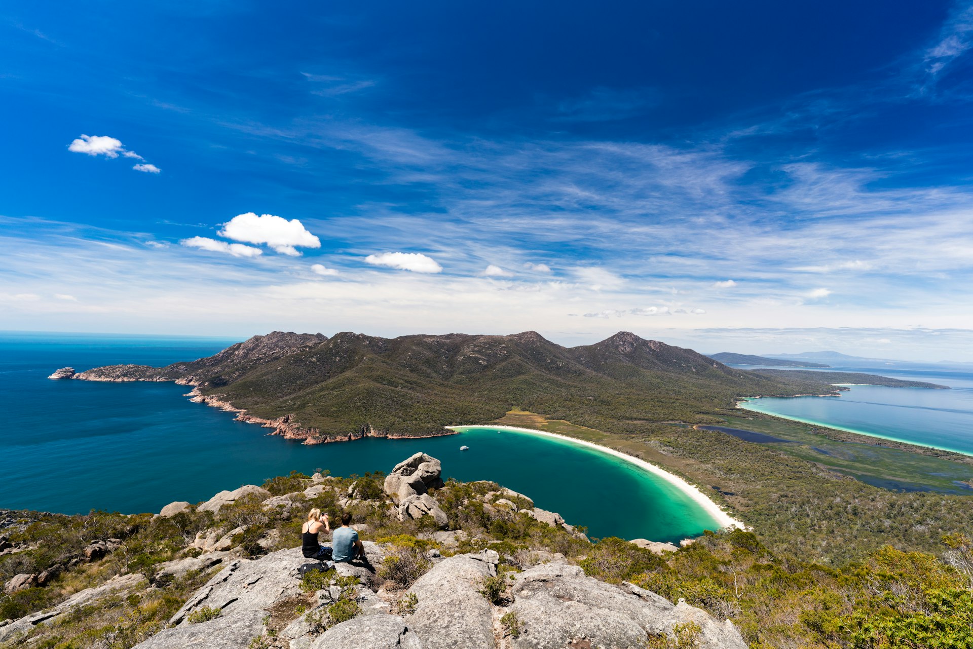 An aerial view of two people on top of Mt Amos overlooking Wineglass Bay in Tasmania