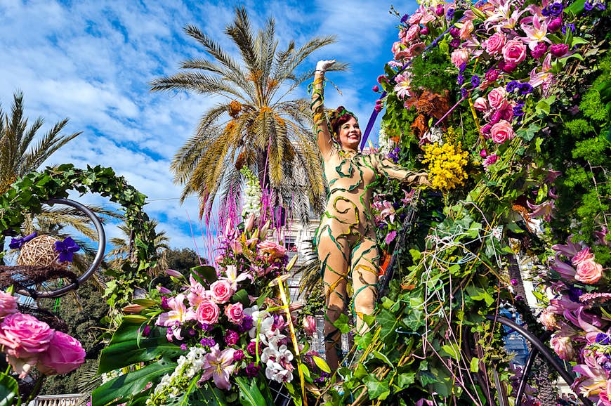 A woman in a nude body suit waves at crowds from a parade float covered in flowers