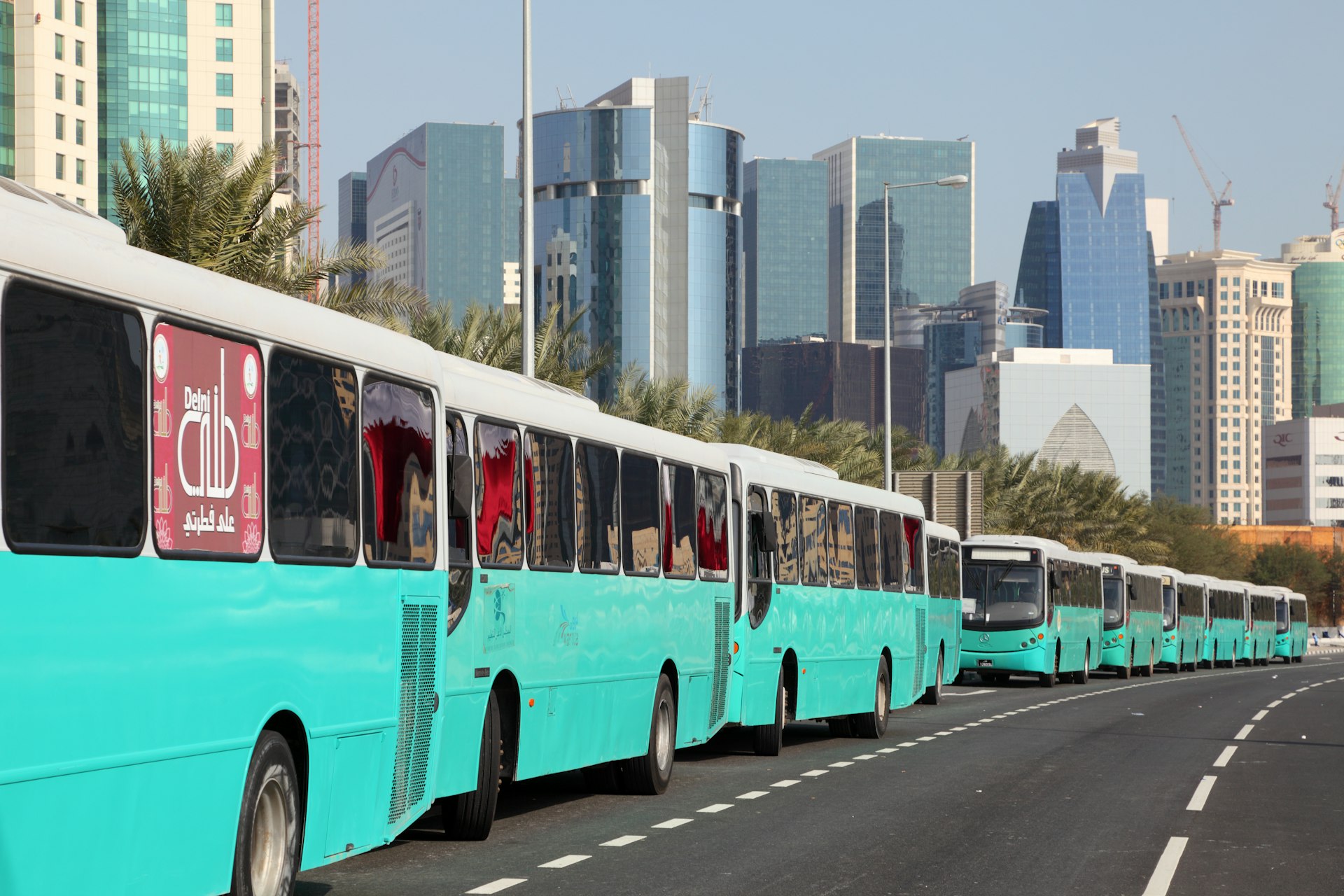 Several turquoise blue buses are lined up on a road with towers seen beyond in Doha, Qatar