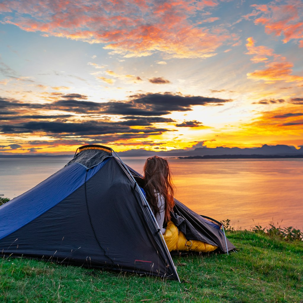 Woman watching the morning sunrise over water from her tent while wild camping in the Isle of Skye, Scotland, UK; Shutterstock ID 1822498943; your: Tasmin Waby; gl: 65050; netsuite: Online Editorial; full: Demand Project