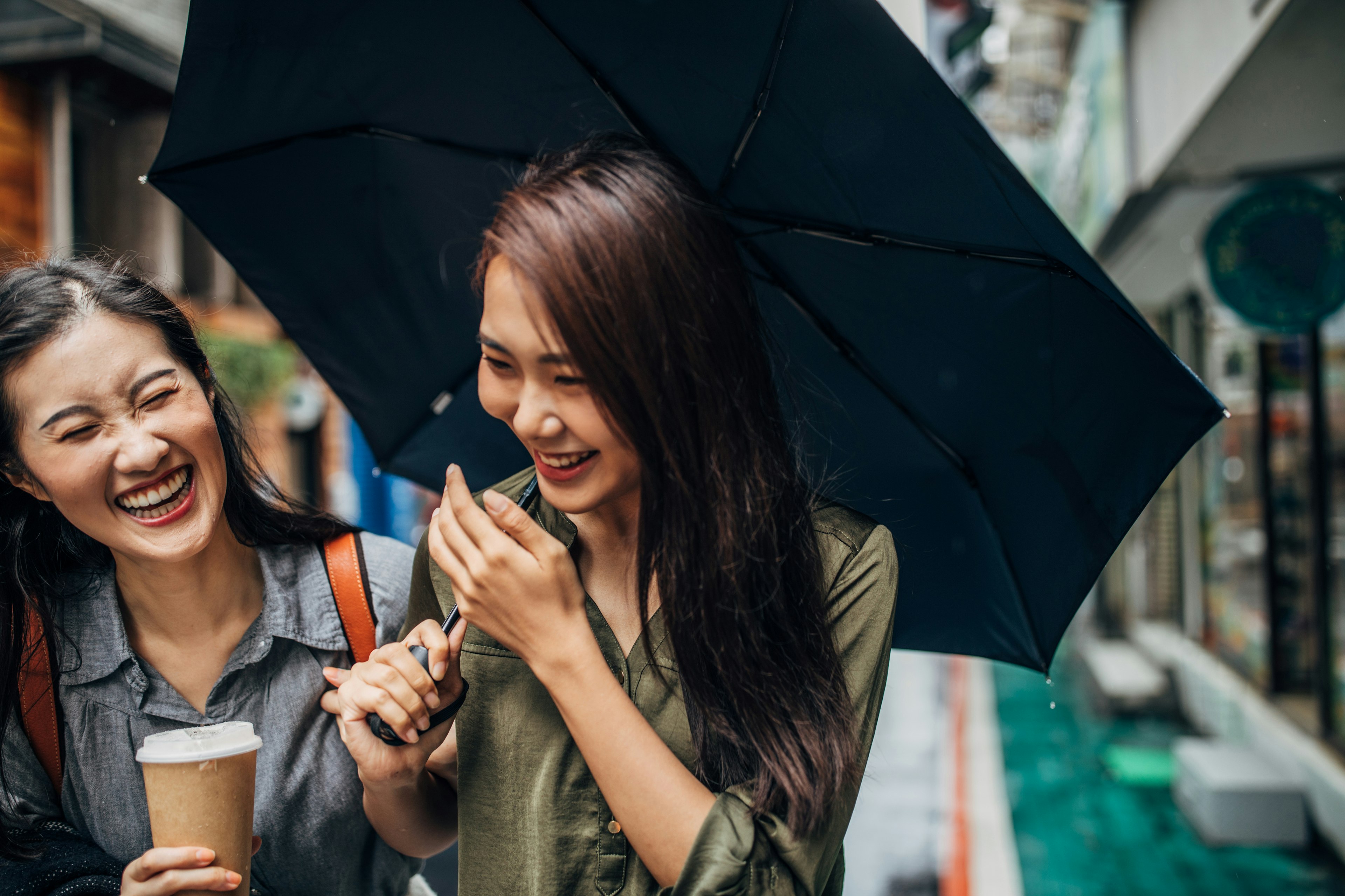 Two cheerful female friends walking on the street, holding umbrella and takeaway coffee, talking and laughing.