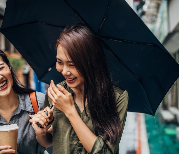 Two cheerful female friends walking on the street, holding umbrella and takeaway coffee, talking and laughing.