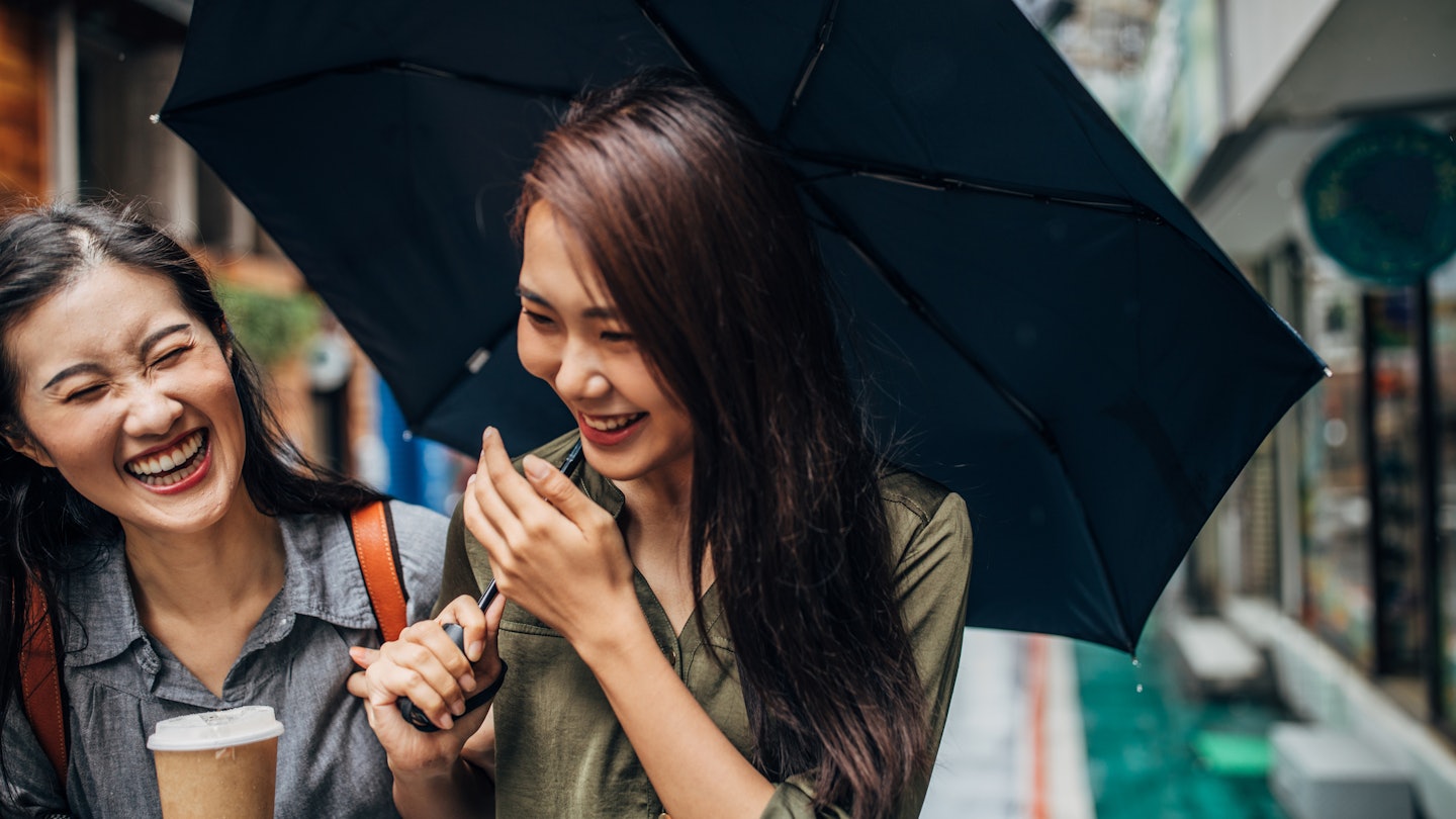 Two cheerful female friends walking on the street, holding umbrella and takeaway coffee, talking and laughing.