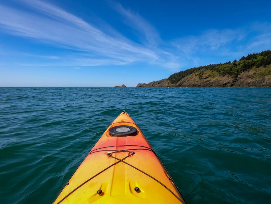L'avant d'un kayak jaune-orange dans des eaux d'un bleu profond avec le littoral devant