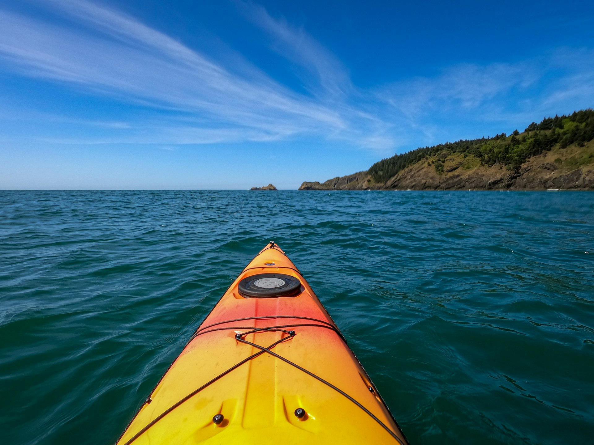 The front of a yellow-and-orange kayak in deep blue waters with the coastline ahead