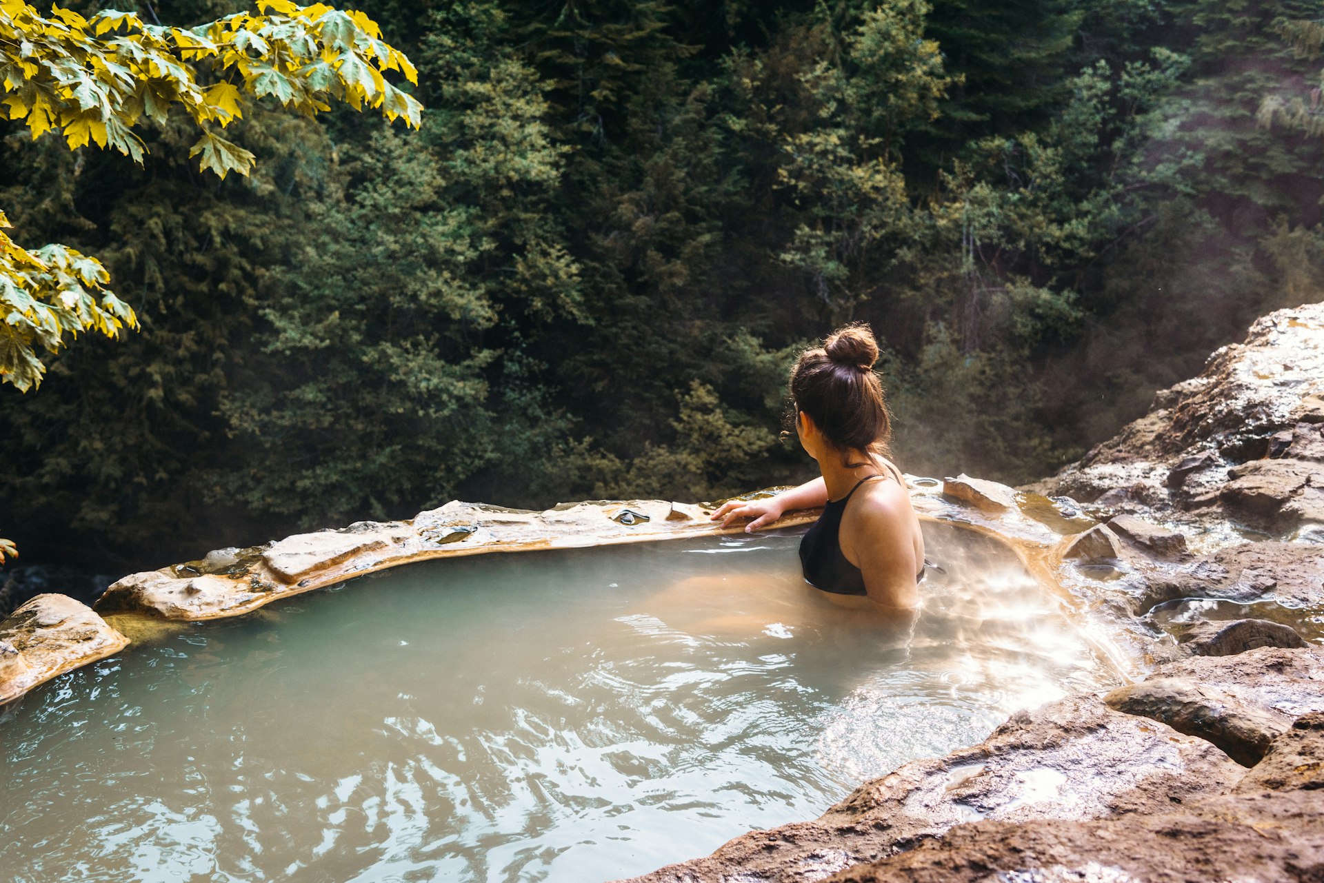 A woman sits in a hot spring surrounded by woodland
