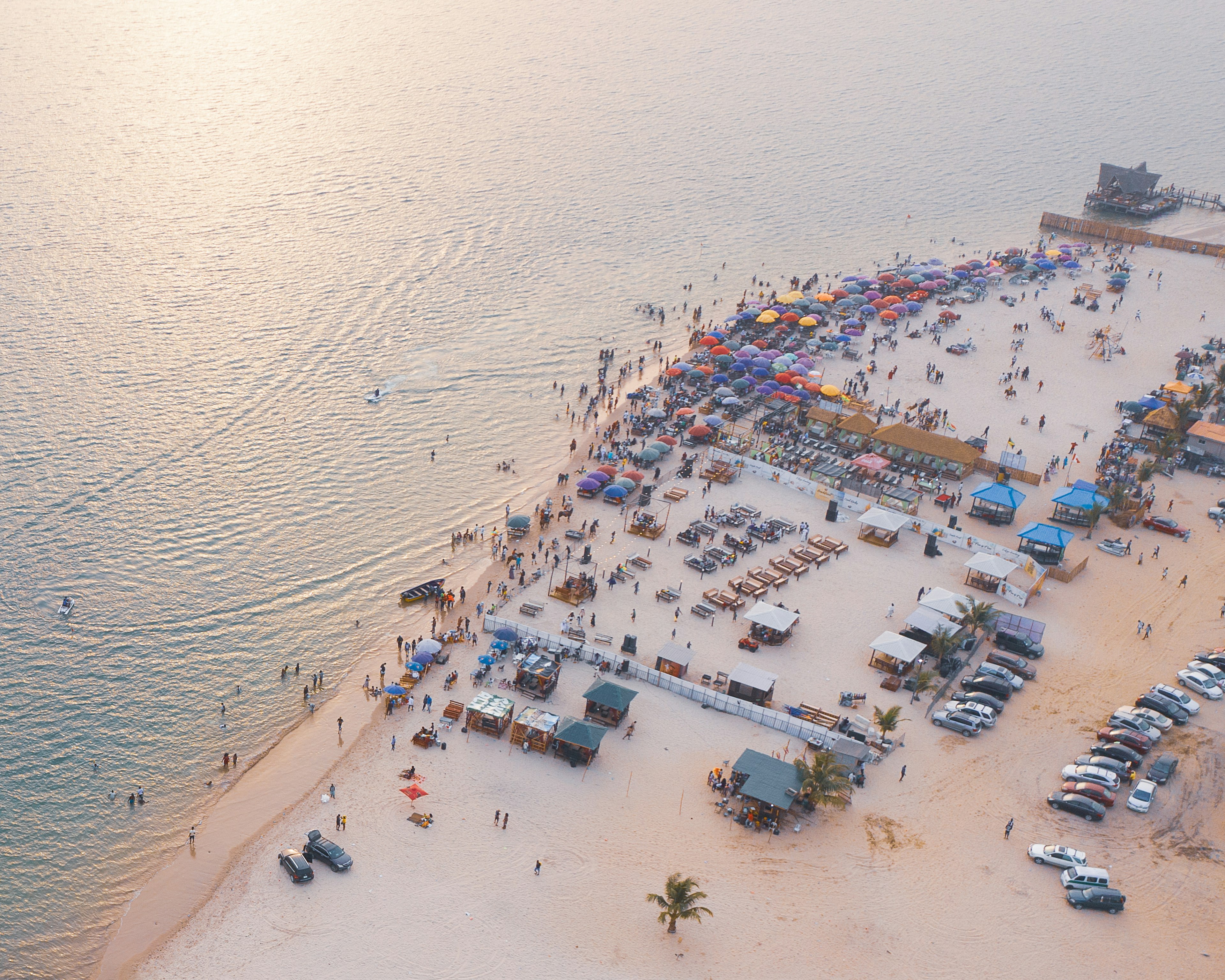 An aerial view of a vast sandy beach with lots of colorful umbrellas right at the water's edge as the setting sun leaves an orange glow in the sky