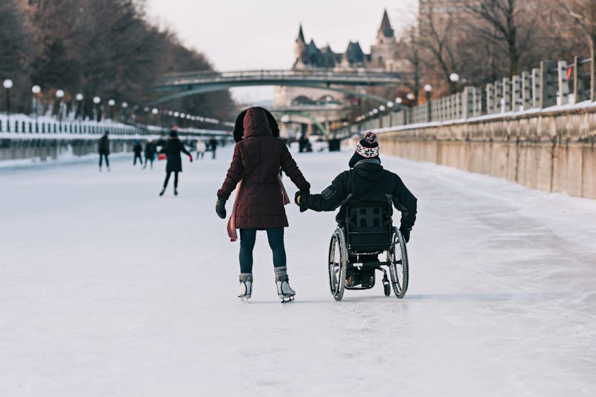 Two people on a frozen canal holding hands; one is on ice-skates, the other is in a wheelchair