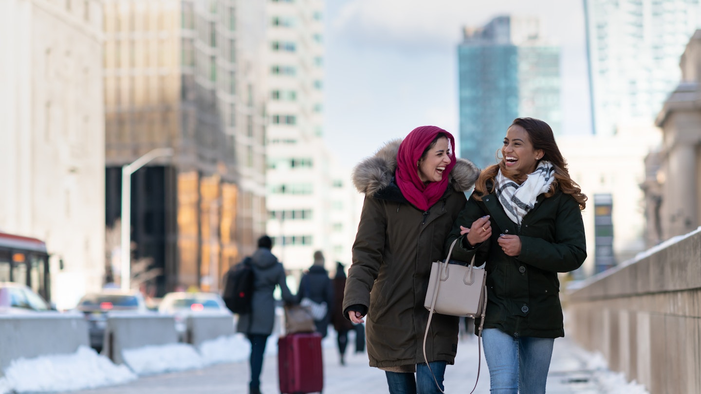 Two (2) attractive muslim adult female friends smile as they walk through the city. They are stylish and enjoying a cold winter stroll together.