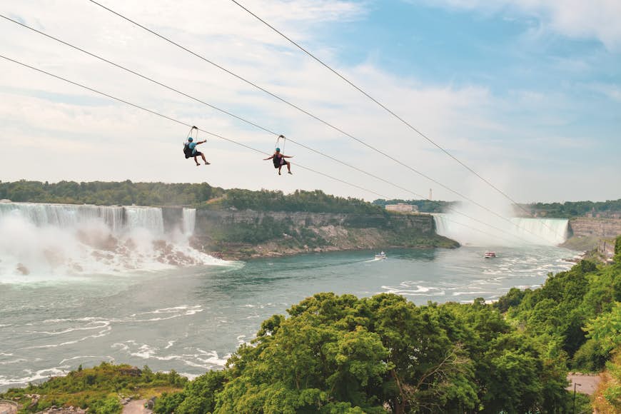 Two zipliners head down a wire towards a vast cascading waterfall