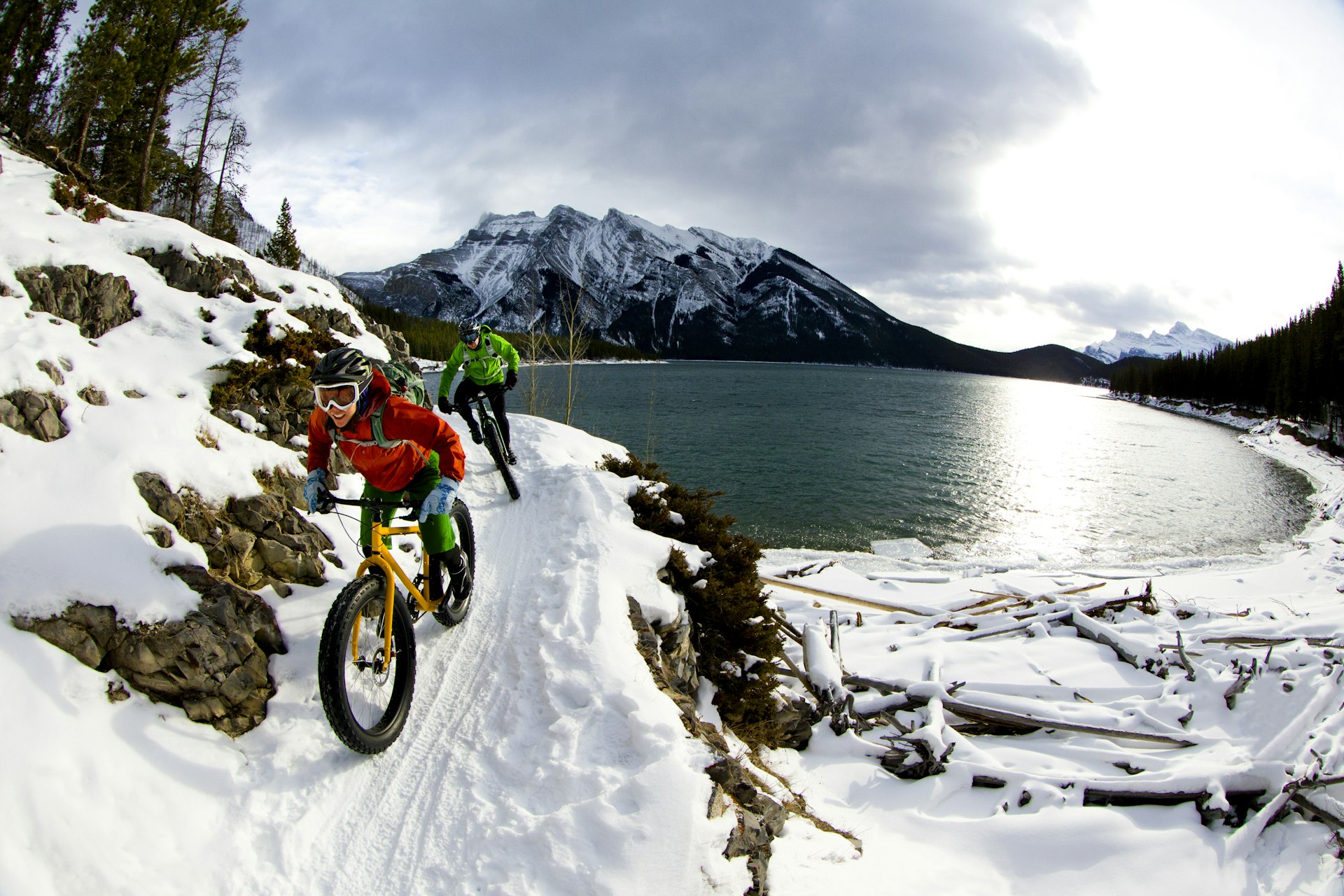 A couple ride bikes with fat tires over a snowy track by a lake in a moutainous area