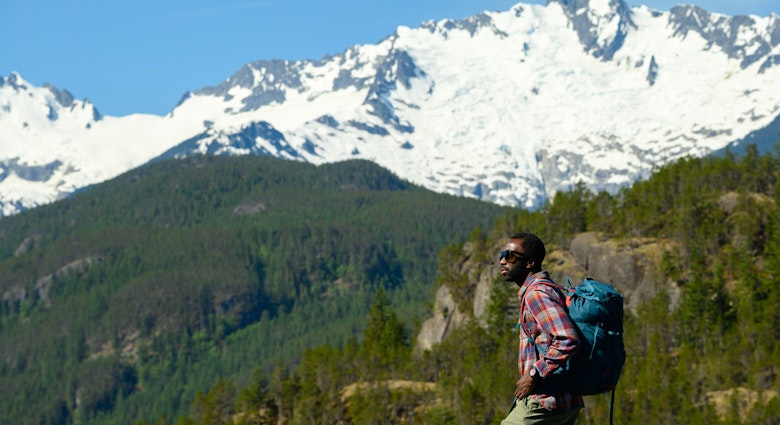 Male hiker looking away while standing against mountains. Mid adult man is spending vacation in forest. He is wearing sunglasses and casuals.