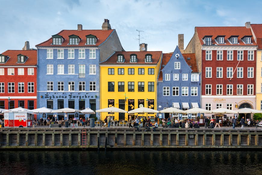 Pastel-colored houses lining a waterway in Copenhagen. People sit at cafes under umbrellas along the waterfront