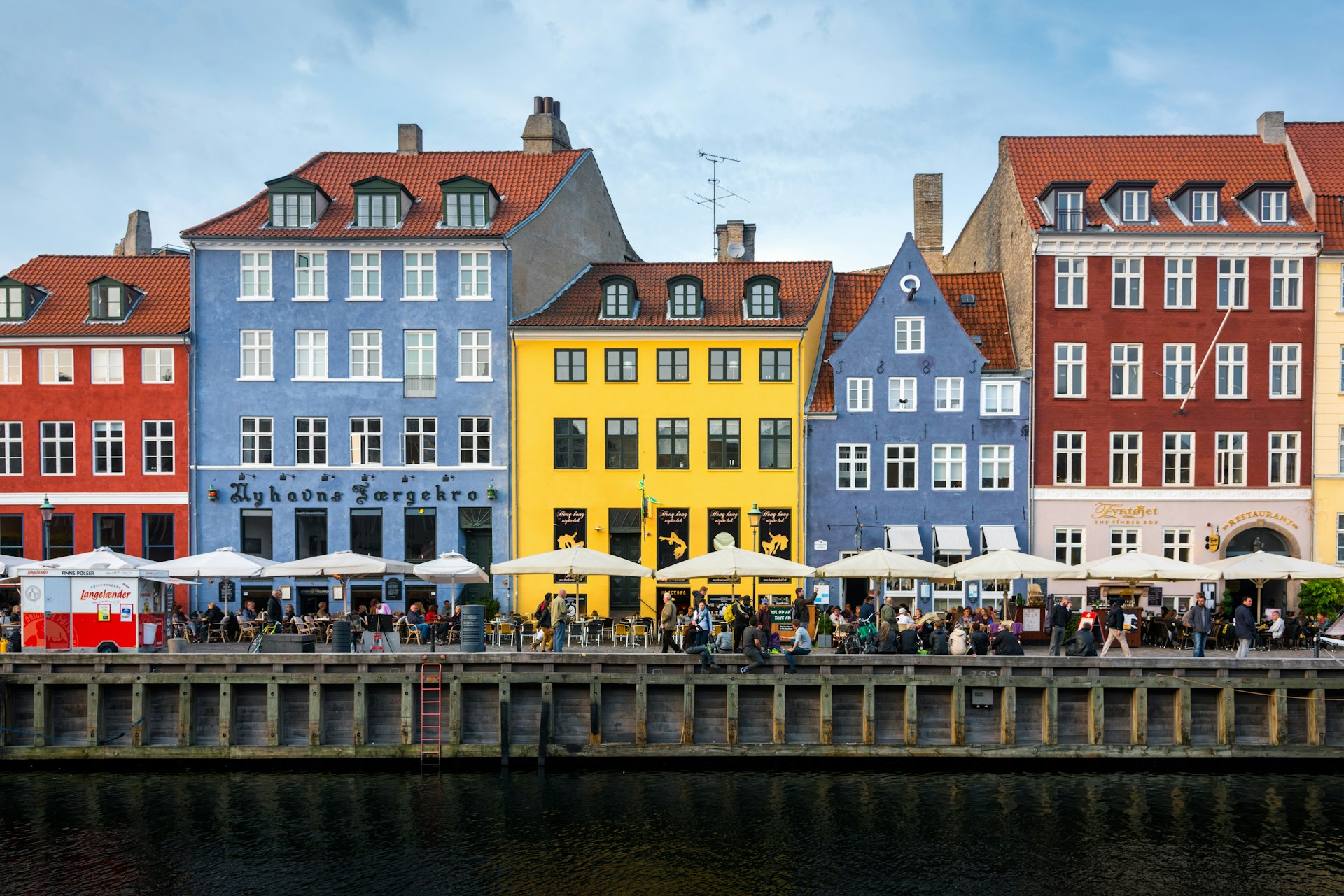 Pastel-colored houses lining a waterway in Copenhagen. People sit at cafes under umbrellas along the waterfront