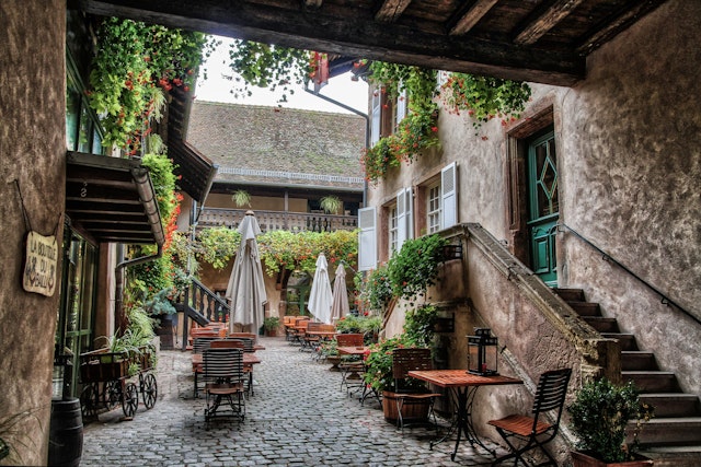 A courtyard of a large stone building covered in greenery with chairs and tables