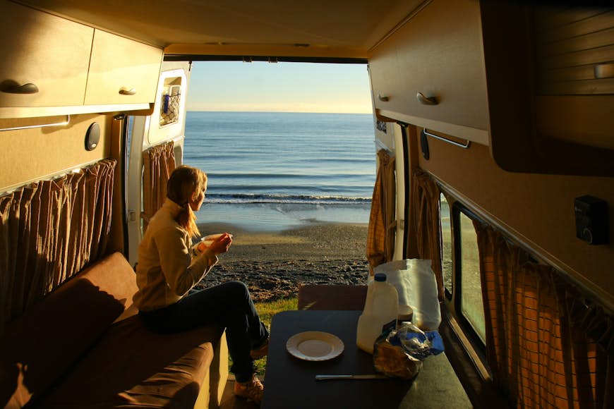 A woman sits in the open doorway of a campervan with a cup of coffee looking out at a sea view