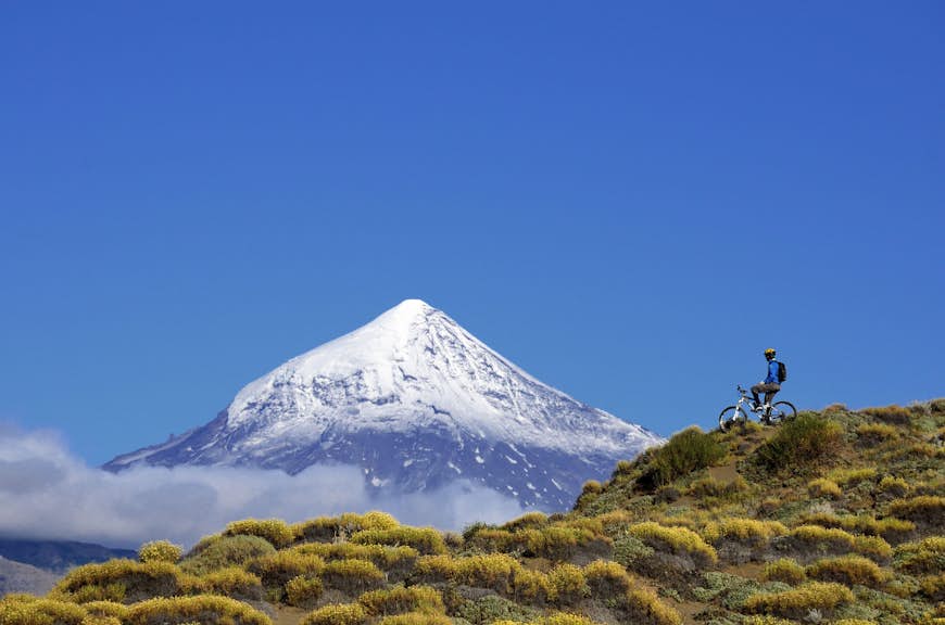 A mountain biker pauses on a hillside and looks toward a distant snow-capped mountain in Argentina