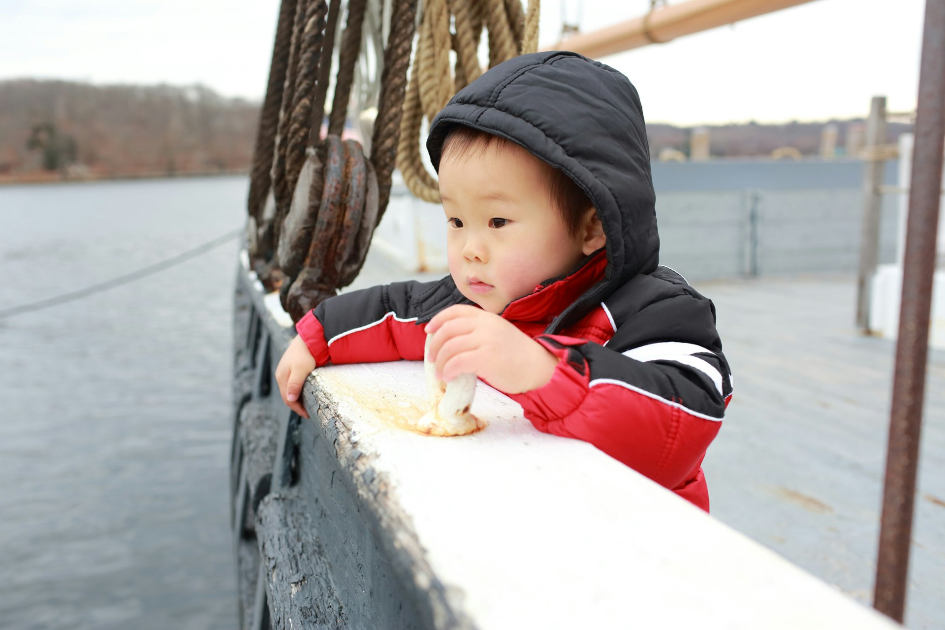 A 20-month old East Asian boy is wrapped in a hat and winter coat and stands looking out at sea from a tall ship in Boston Harbor