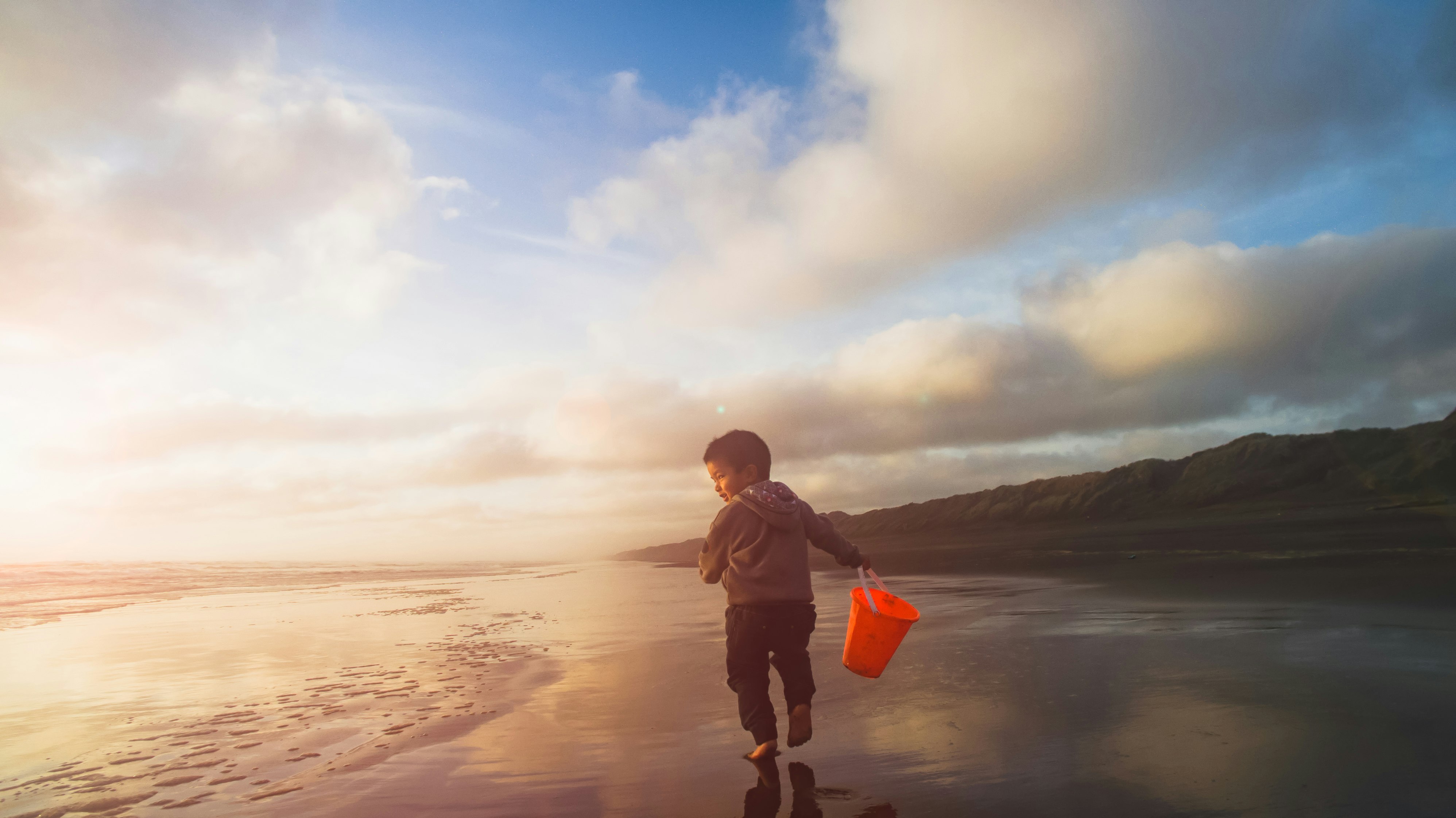 A young child runs a long an empty beach near Auckland holding a red bucket at sunset  