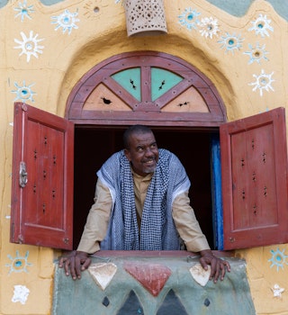 Restaurant owner peering out of window in traditional Nubian Galabeya dress in rural Nubian Village, Aswan Egypt.