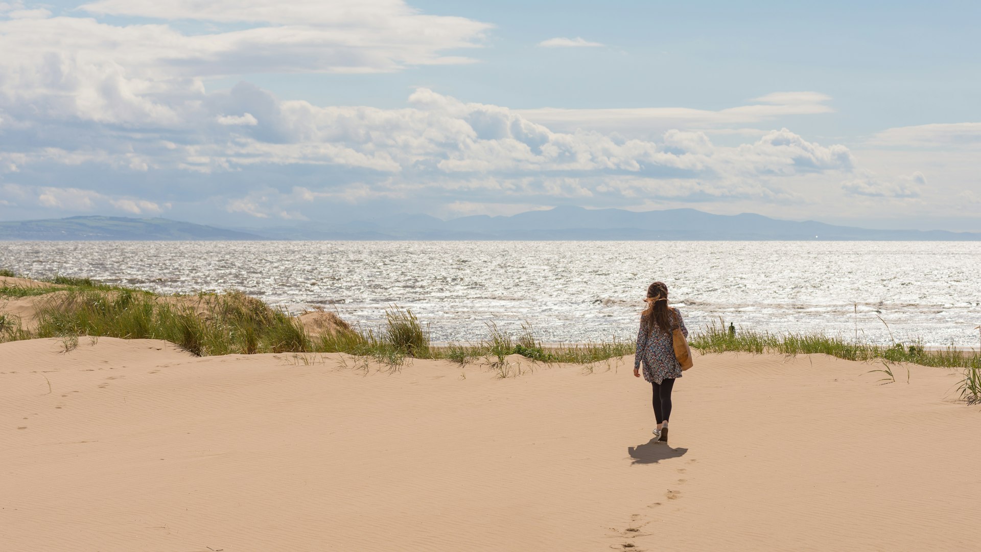 A woman walks on a sand dune to get a view of the north see in the background
