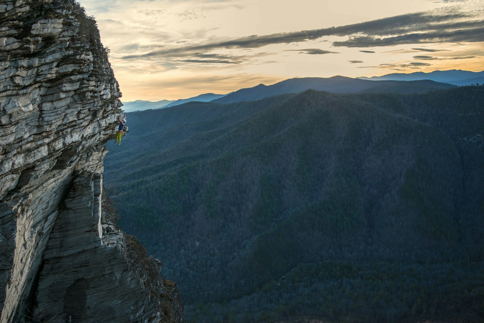 Distant View Of Man Rock Climbing Against Mountains During Sunset