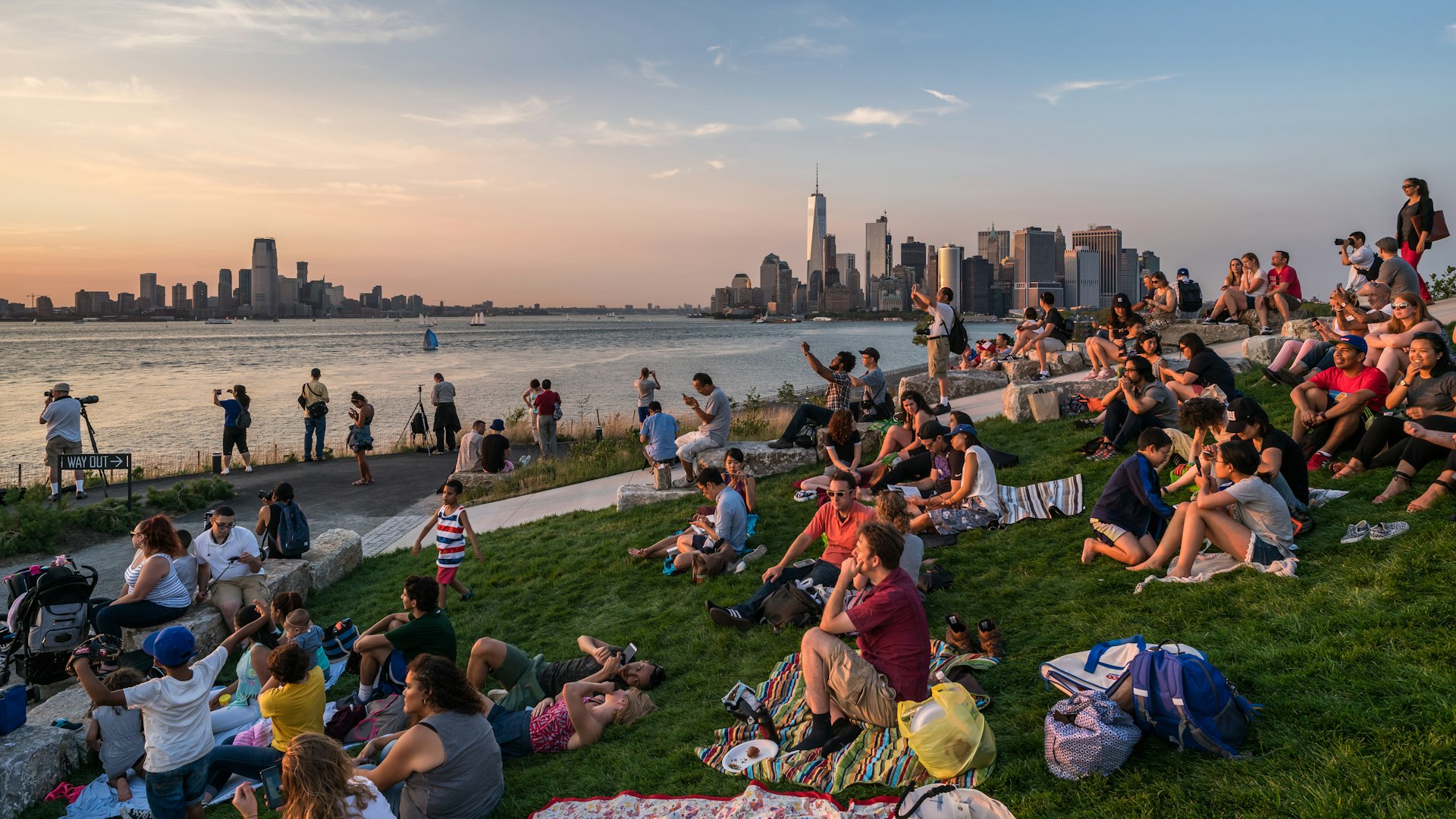 A crowd of people sitting on blankets watch the sunset from Governors Island with the New York City skyline in the background