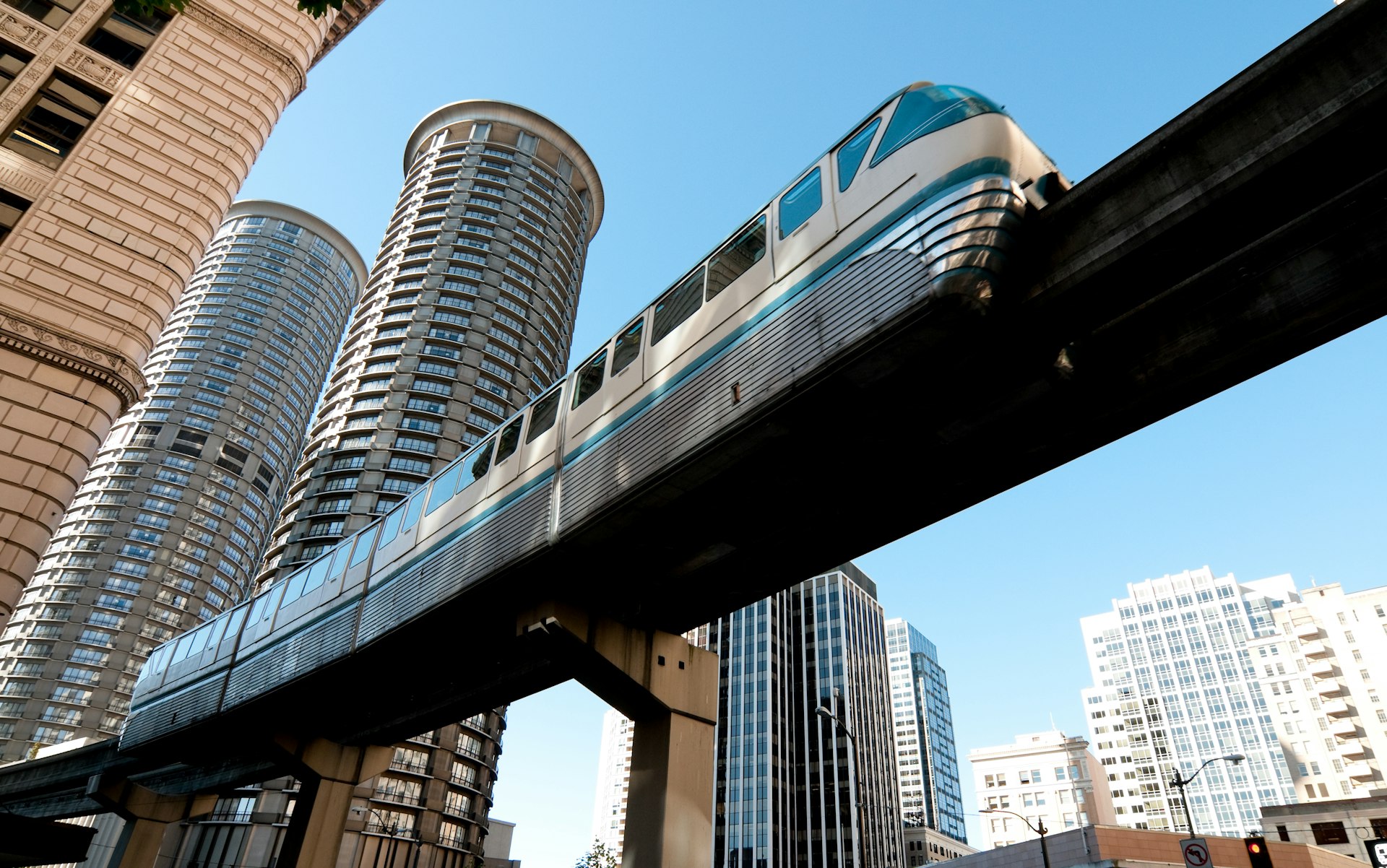 A monorail train runs overhead in a city with tall glass and steel buildings