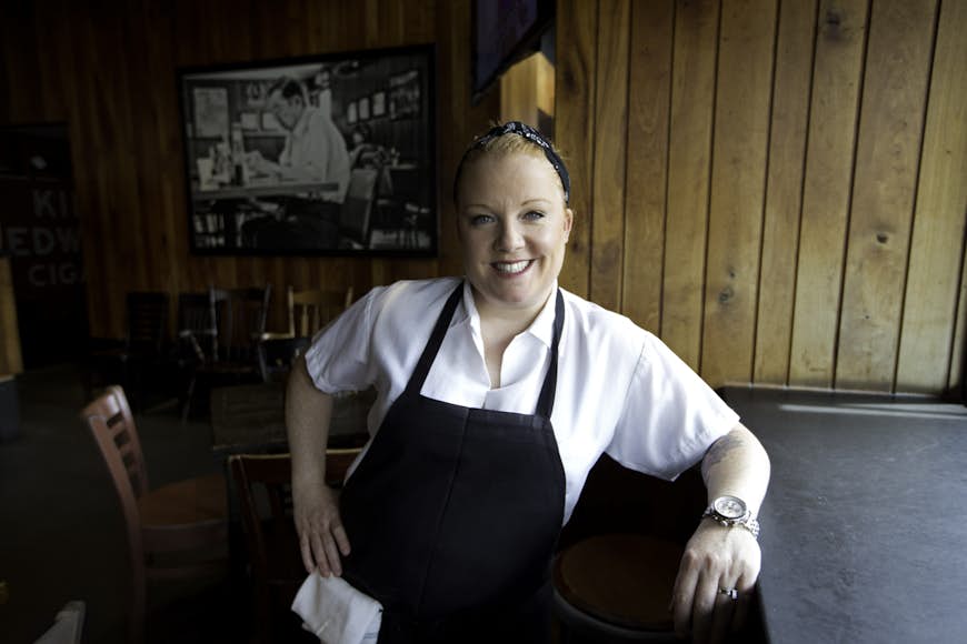 A woman wearing a white shirt and black apron in a restaurant setting smiles at the camera 