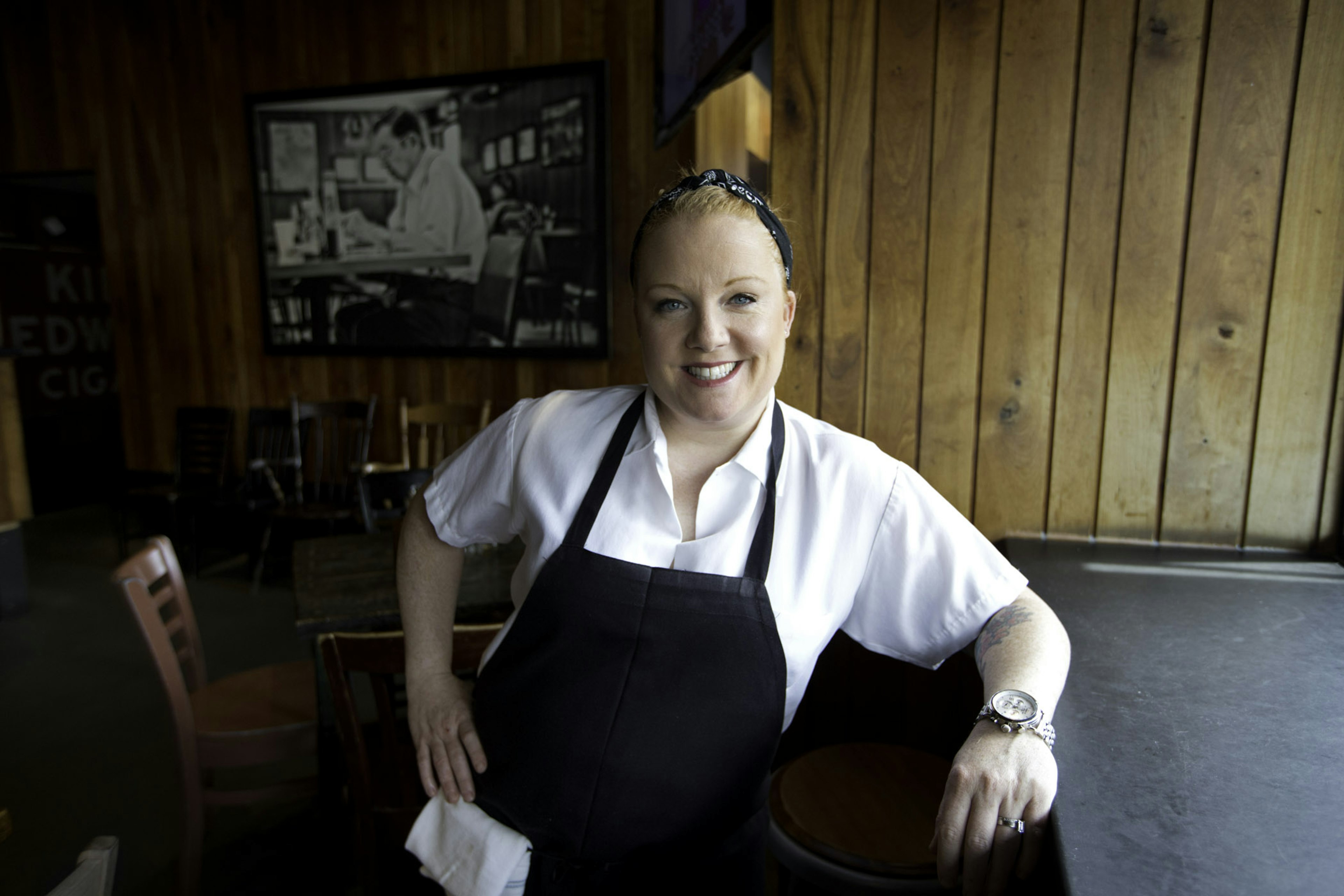 A woman wearing a white shirt and black apron in a restaurant setting smiles at the camera