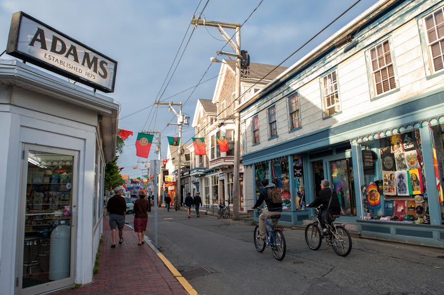 Two cyclists pedal down a shop-lined street in Provincetown