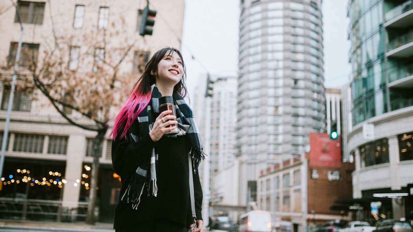 A young woman enjoys time in the city of Seattle, Washington.  She walks the streets, holding a reusable coffee container.
